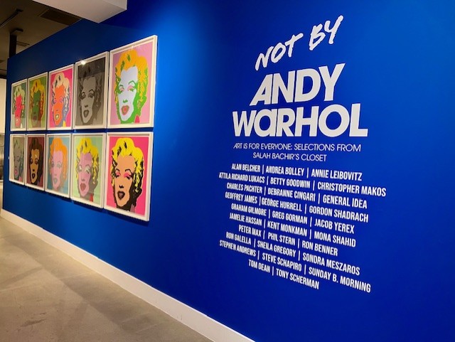 Long view down blue wall with a grid of images of Marilyn Monroe . White lettering reads: Not by Andy Warhol, Art is for Everyone: Selections from Salah Bachir's closet.