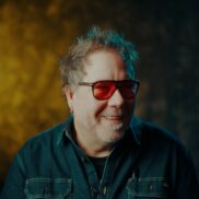 man with messy hair and red sunglasses smiles and looks beyond the camera wearing a denim shirt